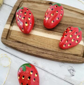 4 painted strawberry rocks, 3 on a wood cutting board and one beside them on a table top