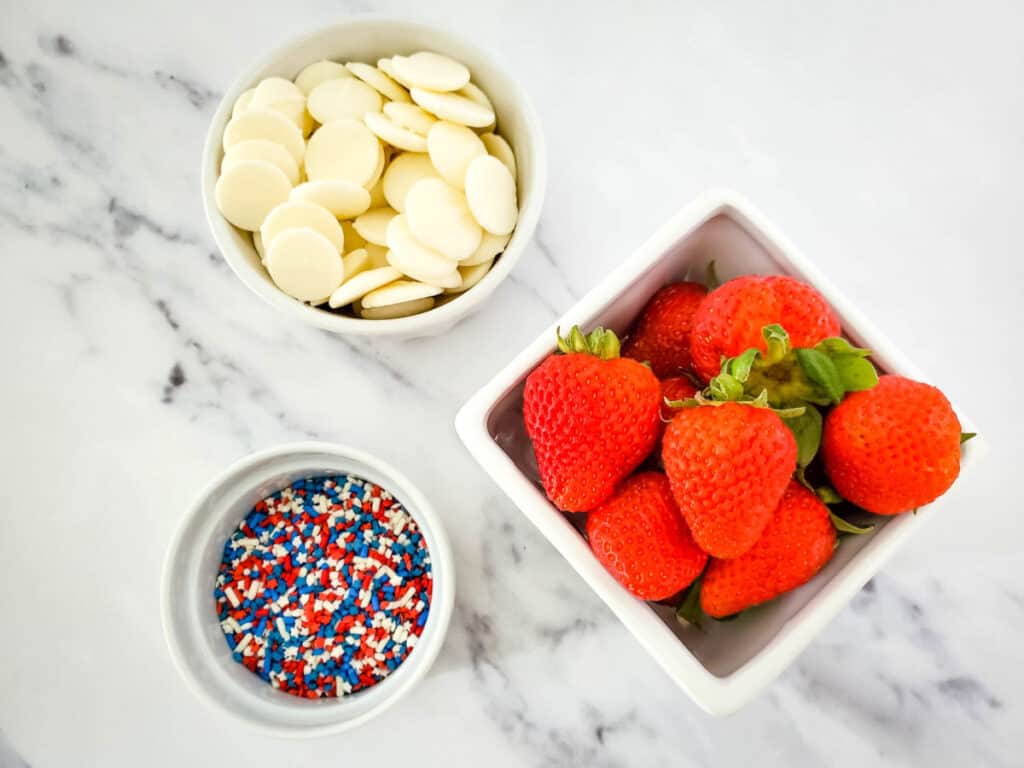 ingredients for patriotic white chocolate dipped strawberries, a bowl of white chocolate candy melts, a pint of strawberries and a small bowl of starts and stripes sprinkles in red white and blue