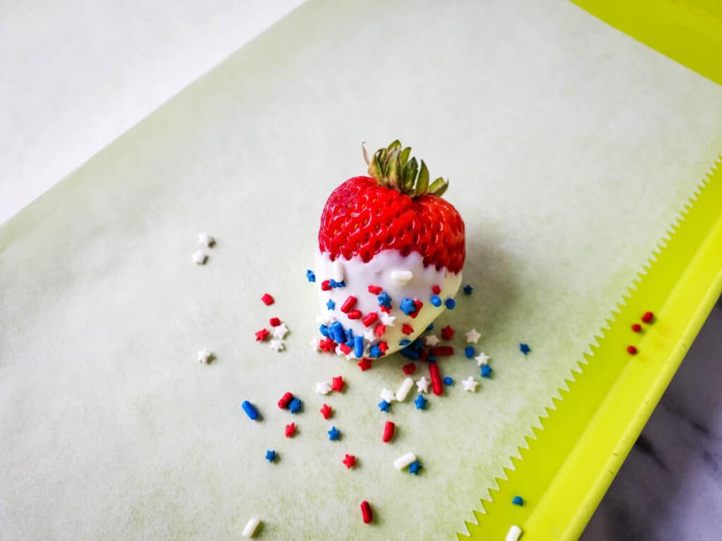 adding sprinkles to a strawberry that was just dipped in melted white chocolate on a cookie sheet lined with parchment paper