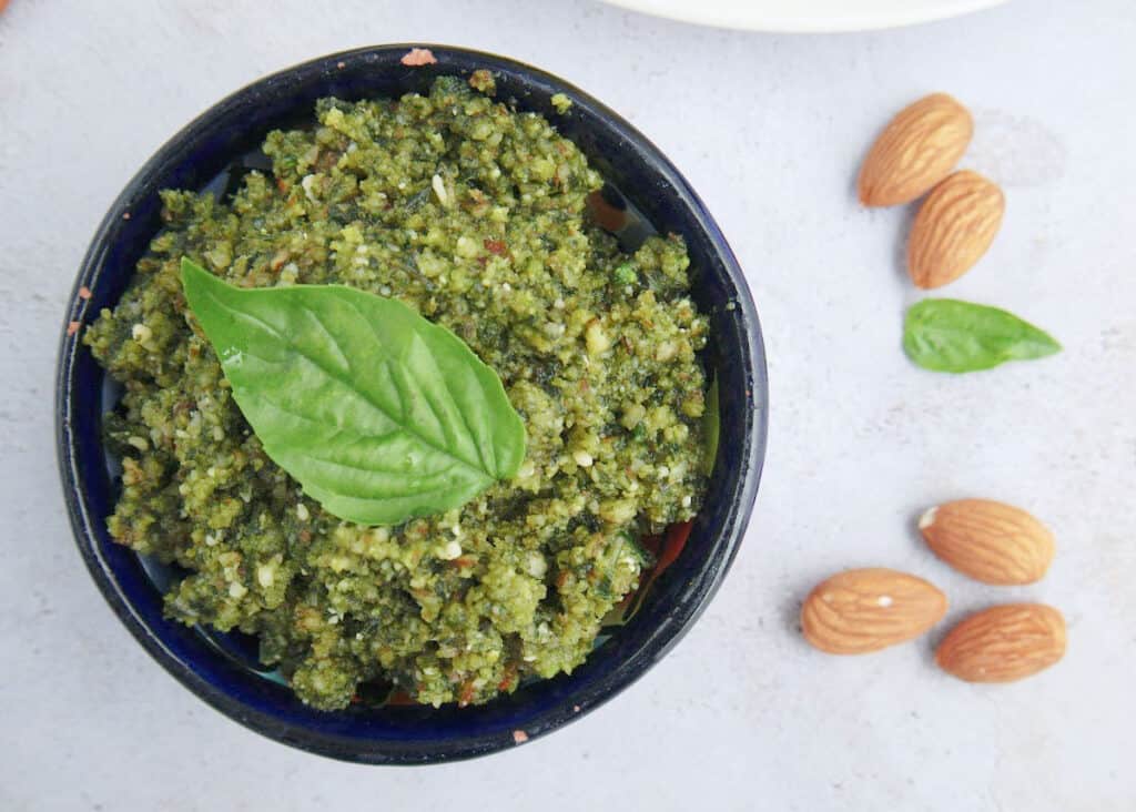 a bowl of basil almond pesto with a basil leaf on top and a few almonds on the table beside it
