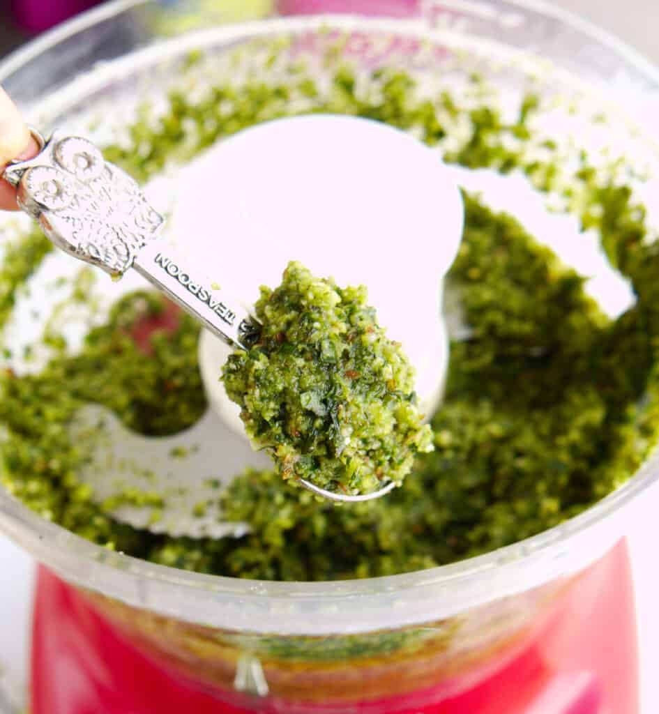 a spoonful of the finished basil almond pesto above the food processor filled with the finished basil almond pesto