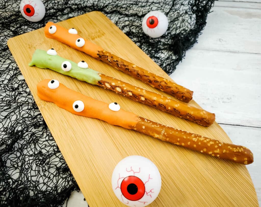 3 finished halloween monster pretzel rods on a cutting board with toy eyeballs