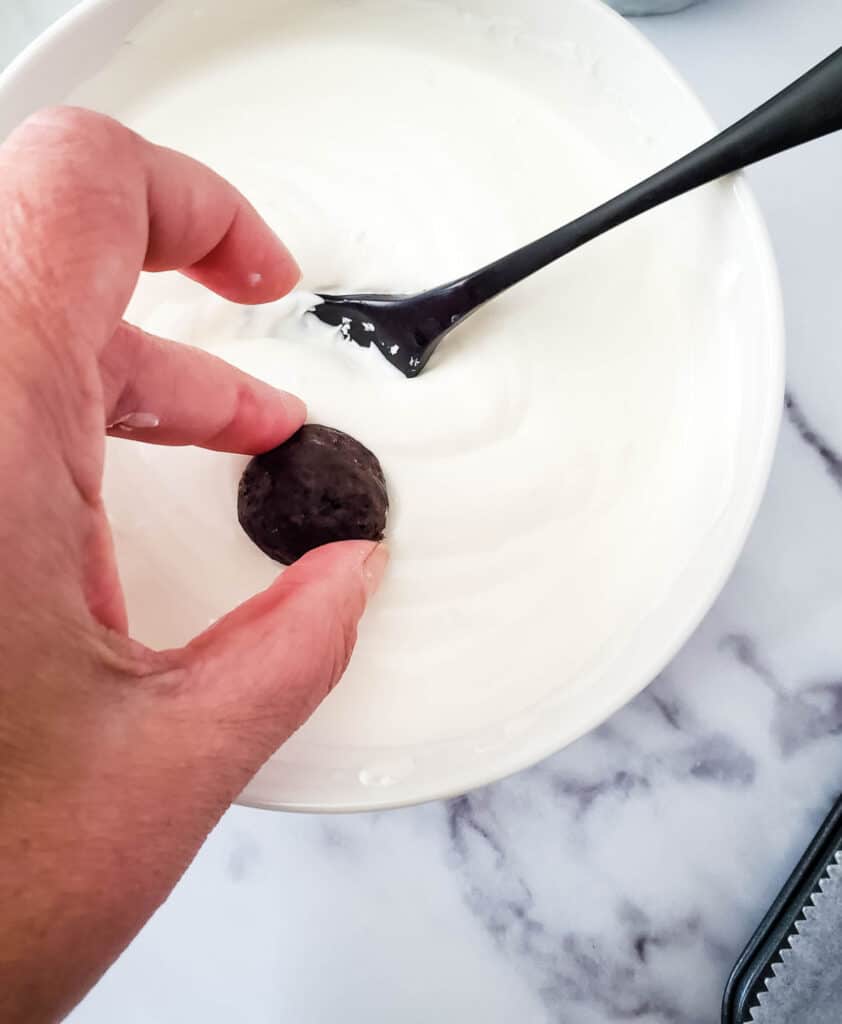 a bowl of melted white chocolate candy melts, being spooned over the oreo cookie balls