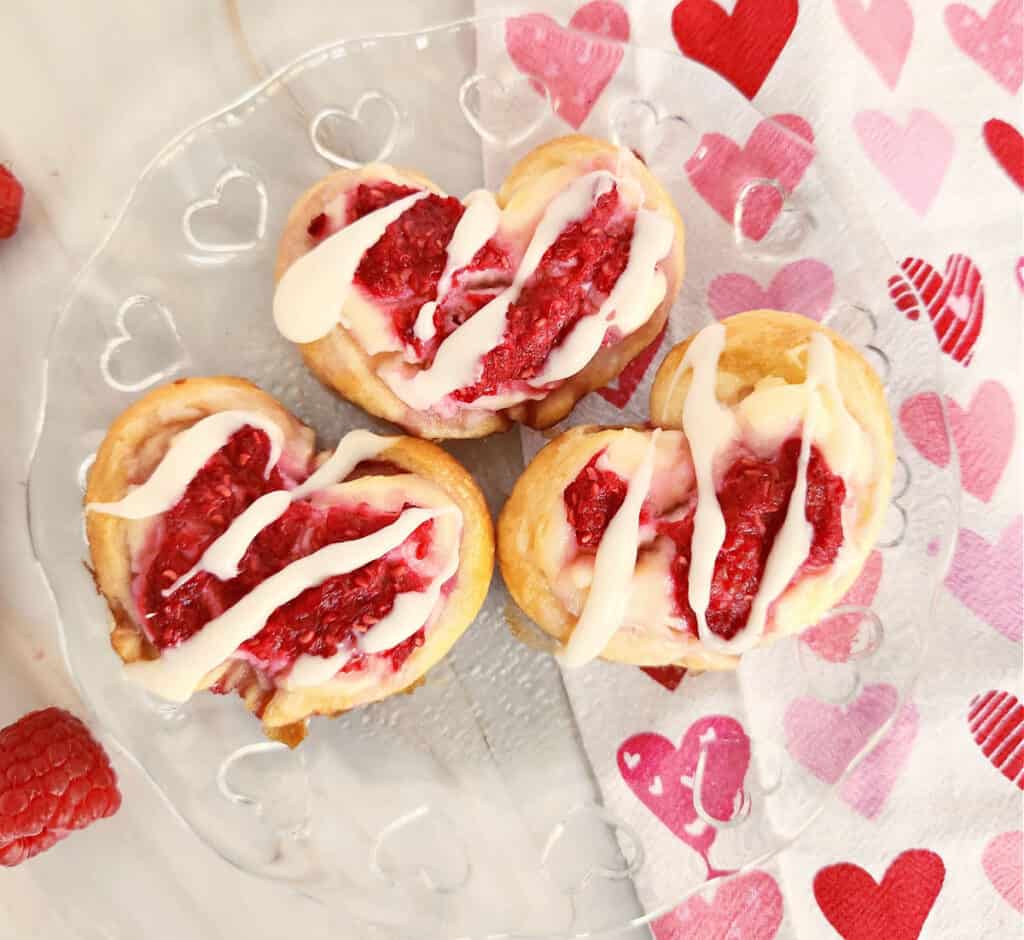 a clear plate with 3 finished heart shaped valentines day raspberry cream cheese danish pastries ready to be served by a napkin with hearts on it
