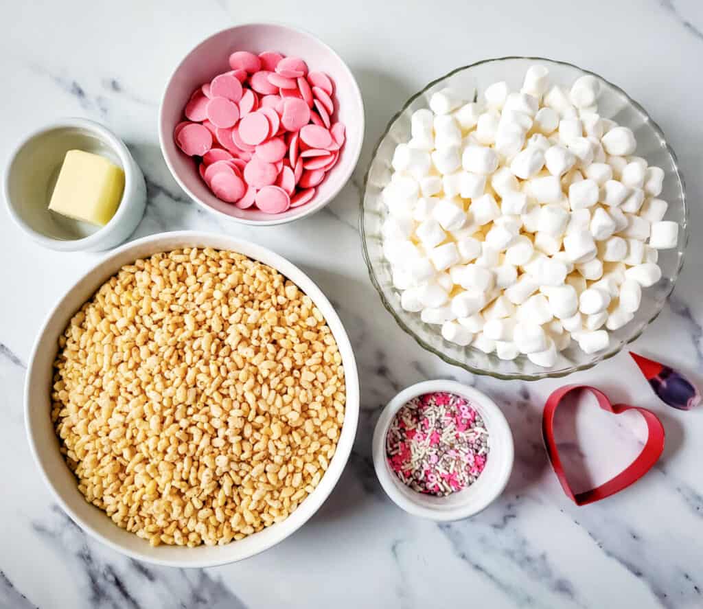 ingredients laid out for Valentines Rice Krispie Treats, butter, candy melts, marshmallows, rice krispies, sprinkles, food coloring and a heart shaped cookie cutter